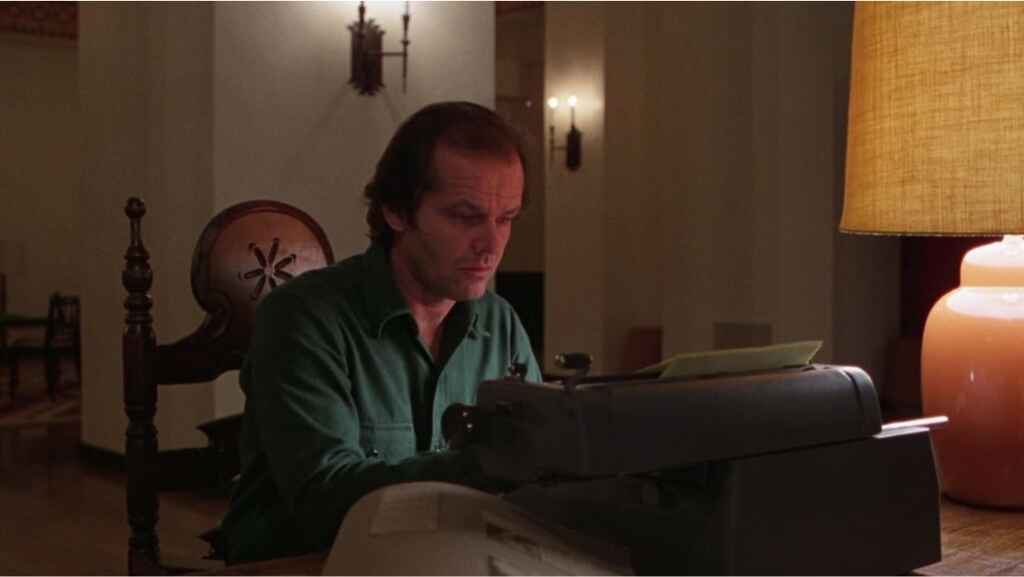 Jack Nicholson as Jack Torrance in The Shining. He is wearing a green sweater and sitting at a desk working on a typewriter.