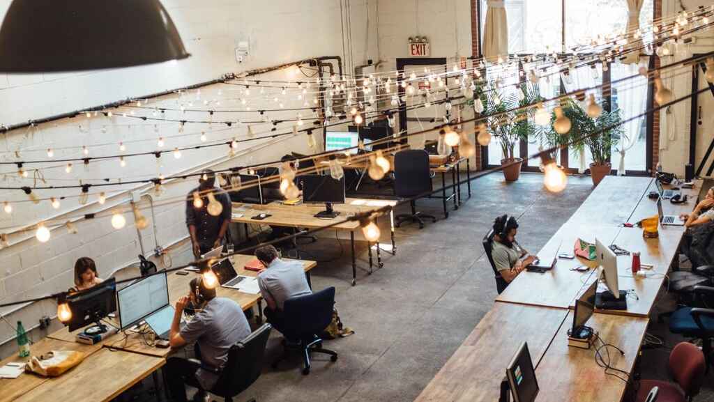 An open plan office seen from a bird's eye view. There are multiple strands of Edison lights hanging from the ceiling. At long light wooden tables multiple people sit working at computers, many of them wearing headphones.