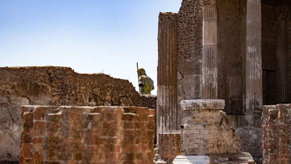 The ruins of pillars and walls with the broken statue of a man in the center set against a bright blue sky.