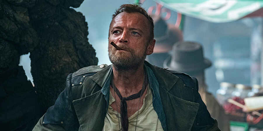 A man wearing a dirty green jacket with a loose tie and open white shirt with a cigar in his mouth