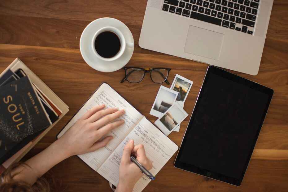 A person writing in a notebook, surrounded by books, a laptop, Polaroids, and a cup of coffee