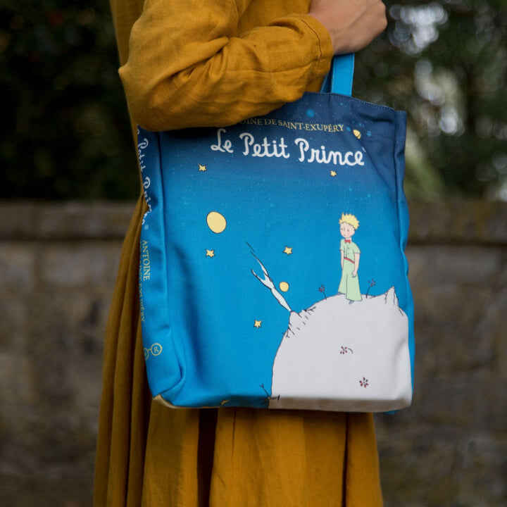 Woman in a yellow dress holding a blue Le Petit Prince tote bag.
