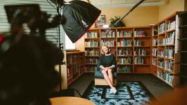 A woman smiles in an armchair in front of a bookcase. In front of her can be seen microphones, an overhead light, and a camera used for filming an interview.