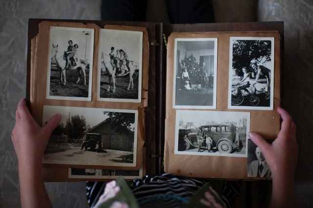 Two hands hold an aged photo album open. Inside can be seen 4 black and white photos of people walking, riding horses, and smiling to the camera.